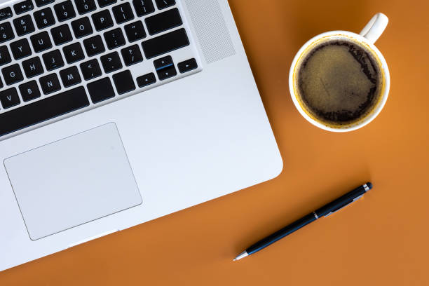 Laptop, cup of coffee and pen on a brown background, flat lay. Minimalistic office desk, top view.