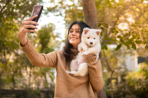 Outdoor portrait of beautiful happy young woman using smartphone to take selfie with her cute pet dog while having fun in the park at day time.