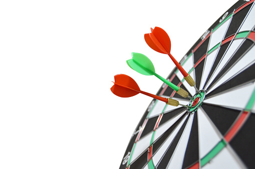 Cutout of three darts, two red and one green hitting bull's eye spot on on a tilted dart board isolated over white background denoting success, win, achievement of business target, competition