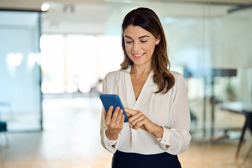 Mid aged mature happy professional business woman using mobile applications on phone at work standing at lobby, businesswoman entrepreneur holding smartphone reading messages on cellphone in office.