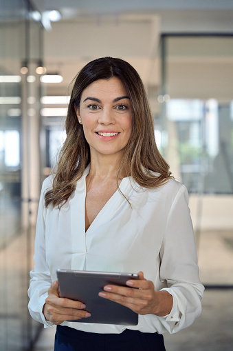 Smiling mid aged mature professional business woman company manager executive wearing white blouse holding fintech tab digital tablet standing in office at work, looking at camera, vertical portrait.