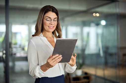 Smiling mid aged mature professional business woman banking manager, 40s female executive or entrepreneur holding fintech tab digital tablet standing in office at work, looking at pad.