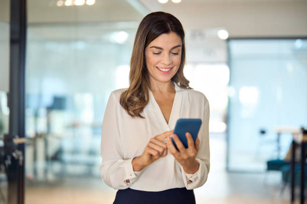 istock Smiling mid aged business woman executive using smartphone standing in office. 2004891085