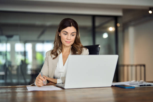istock Busy mid aged business woman working in office using laptop writing notes. 2004891094