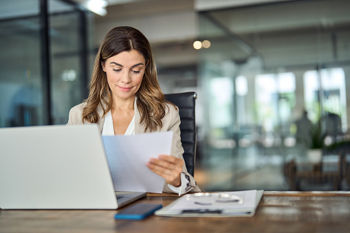 Busy mid aged business woman working in office with laptop reading document. Mature professional female manager lawyer attorney holding paper finance report sitting at desk in office. Copy space
