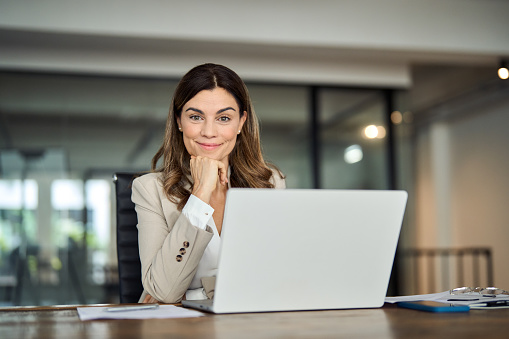 Smiling middle aged 40s professional business woman in office, portrait. Happy mature mid age professional businesswoman executive wearing suit looking at camera at work with laptop sitting at desk.
