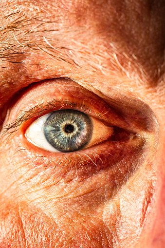 Color image depicting an extreme close up macro view of the eye of a senior man in his 70s. The man has blue eyes and they are a little bit bloodshot with the capillaries visible.