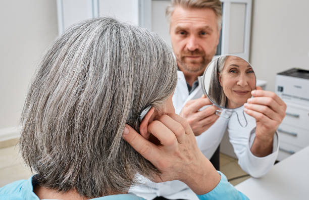 istock Senior woman trying on a hearing aid while looking into a mirror held by a hearing aid specialist in audiology clinic. Stylish hearing device 2005000555