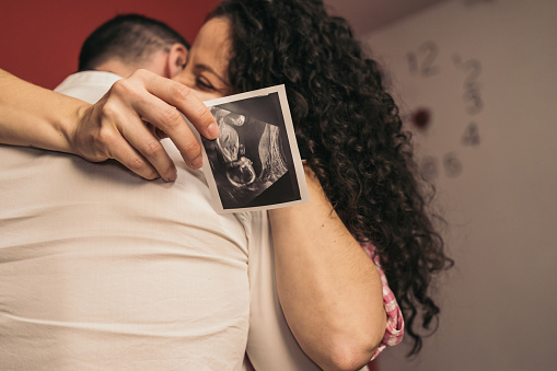 Pregnant Woman Embracing Future Baby’s Father Holding Printed Ultrasound. Young pregnant girl hugging partner holding baby’s ultrasound print.