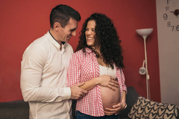 young pregnant heterosexual couple embracing and sharing a loving smile in their living room. a tender moment between a young pregnant couple at home. - human pregnancy couple prenatal care heterosexual couple imagens e fotografias de stock