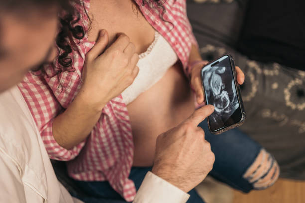 istock Faceless Pregnant Couple on Sofa Watching And Pointing a Phone with Ultrasound Image. Unrecognizable pregnant couple on sofa, woman showing ultrasound image on phone. 2005014840