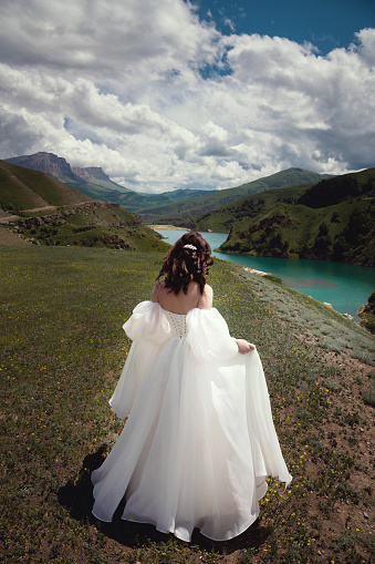 A beautiful bride in a white dress stands or walks in the autumn mountains among green grass against the backdrop of a lake in the distance. Scenic wedding photography.