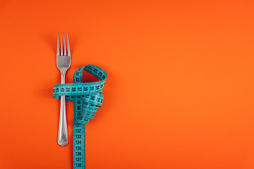 Measuring tape threaded through a fork on an orange background. Good healthy nutrition. Therapeutic fasting. Diet for weight loss concept.