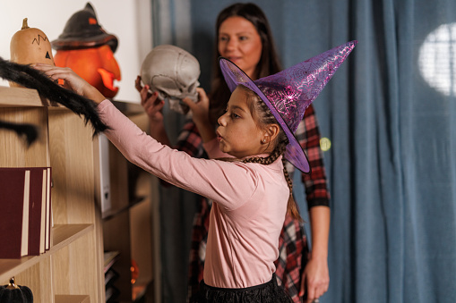 Candid shot of happy young girl and her mother having fun when decorating shelves in their home with various Halloween decorations.