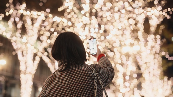 A woman is taking photos and videos of Christmas lights with her mobile smart phone in the city at night.