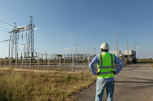 A male engineer in a white helmet and reflective jacket standing on wind turbine power plant, electric power station. Sustainable wind energy concept