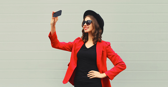 Beautiful stylish lady woman taking selfie with smartphone in red business blazer jacket, black hat