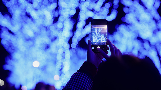 A woman is taking photos and videos of Christmas lights with her mobile smart phone in the city at night.