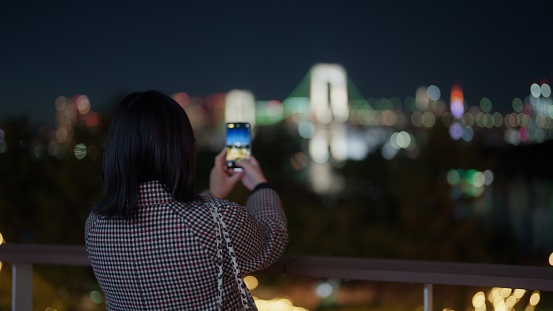 A woman is taking photos and videos of cityscape of Tokyo in Odaiba at night.