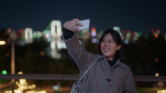 A woman is taking photos and videos of cityscape of Tokyo in Odaiba at night.