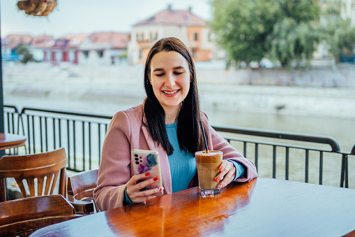 Young cheerful woman using phone while drinking iced coffee at a sidewalk café