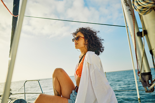 A contemplative young woman lounges on the deck of a sailing yacht, draped in a casual white shirt over her vibrant orange swimwear. Her denim shorts and stylish sunglasses speak to a laid-back day at sea, with her curly hair tousled by the gentle ocean breeze.