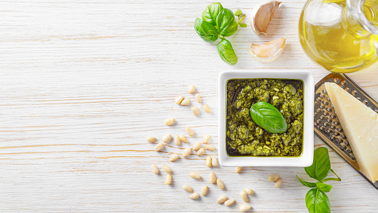 Homemade pesto sauce in small white square jar and ingredients for pasta on white wooden background. Banner, header with copy space. Traditional Italian cuisine, recipe, restaurant menu
