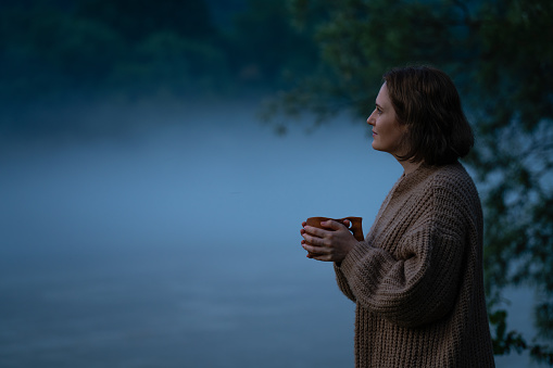 A woman in a sweater on the bank of a foggy river in the evening