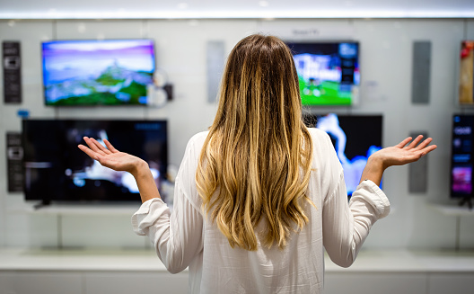 Portrait of young woman having difficulties choosing in store of digital devices. Shopping technology decision people concept