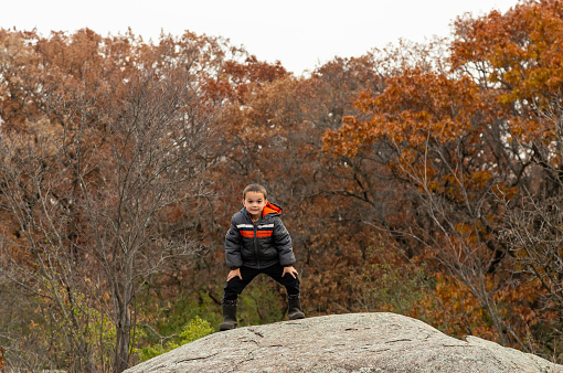 Rear view of a young boy standing at the edge of a large granite rock in the woods on an autumn day.