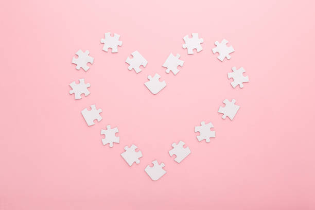 Heart shape created from white puzzle pieces on light pink table background. Pastel color. Closeup. Love and relationship concept. Top down view.