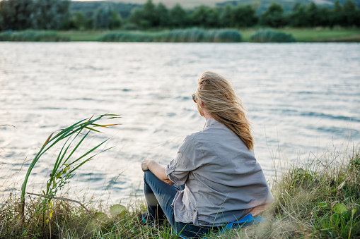 Woman is relaxing in nature by lake to improve her mindfulness and mental health. Enjoyment of fresh air and digital detox outdoors