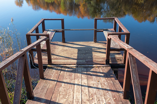 Access to the lake via an old wooden pier. The blue sky and bright autumn trees are reflected in the water. Background.