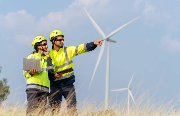 Analyzing Turbine Performance Remotely. Expert engineer in safety suit with team holding a laptop and remote control to access a wind turbine system remotely. Analyzing performance data to optimize energy generation.