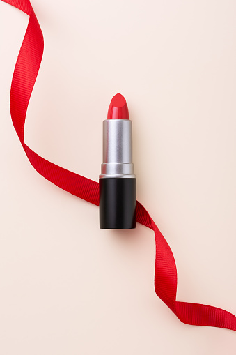 Red lipstick in black and metal tube packaging with red decorated ribbon on beige background. Makeup and cosmetics mock-up product concept.