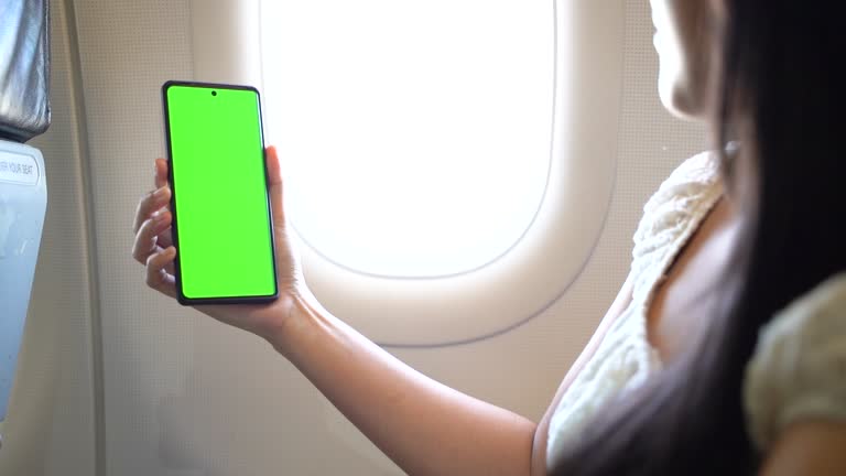 Asian woman holding green screen smart phone in airplane cabin. Successful business woman is flying in an airplane and using a smartphone near the porthole.