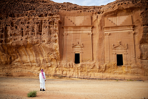 Full length rear view of man in traditional clothing standing in front of entrances to ancient rock-cut burial sites built into sandstone formations.
