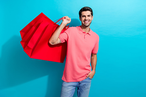 Photo of young handsome guy wearing pink t shirt and jeans holding red packages shopaholic isolated on aquamarine color background.