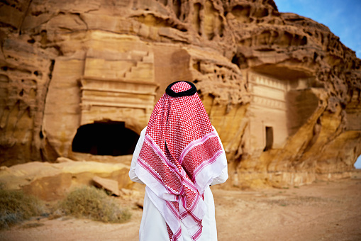 Rear view of visitor in traditional clothing looking at tomb entrances built into natural sandstone formations, erected during Nabataean civilization.