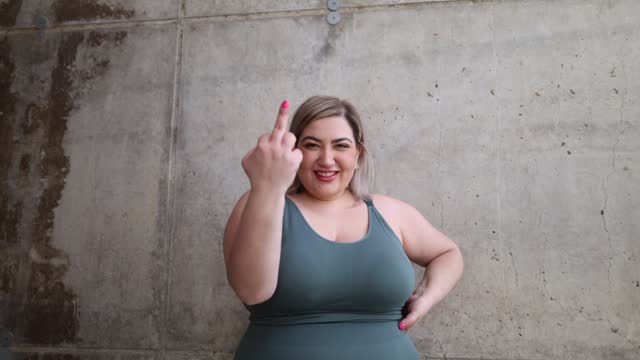 Empowered curvy woman posing showing biceps muscles looking at camera, ends up showing middle finger making bad expression, provocation and rude attitude.