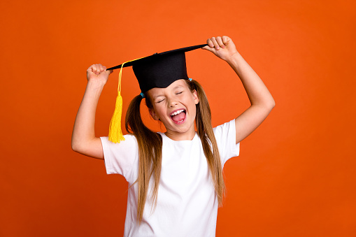 Portrait of overjoyed schoolkid closed eyes hands touch mortarboard hat isolated on orange color background.