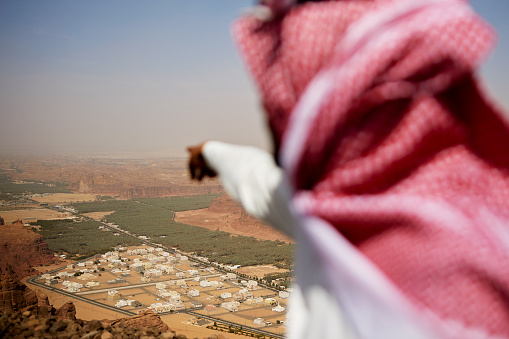 Over the shoulder view with focus on background landscape as Saudi man draws attention to Al-Ula valley, rock formations, canyons, and oasis below.