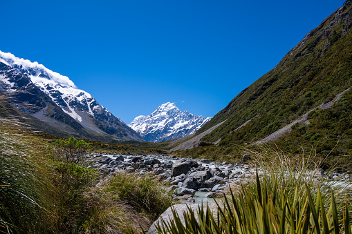Discover the picturesque landscape in New Zealand's South Island leading to the renowned Mount Cook National Park. Behold the stunning sight of snow-capped Mount Cook, majestically framed by other towering mountain peaks. In front a glacier river.