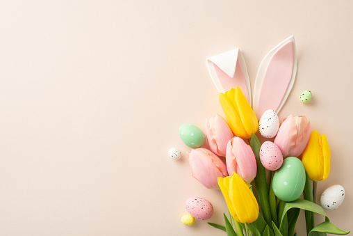 Spring joy captured in top view snapshot of vibrant Easter bouquet, featuring whimsical bunny ears, lively colored eggs, blooming tulips. Pastel beige backdrop offers a perfect canvas for text or ads