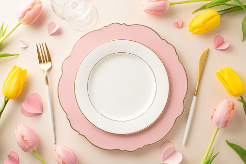 Woman's Day dining arrangement idea. Top view photo of opulent dishware, cutlery, goblet, lovely tulips, and hearts on a creamy beige background, providing space for text or promotion