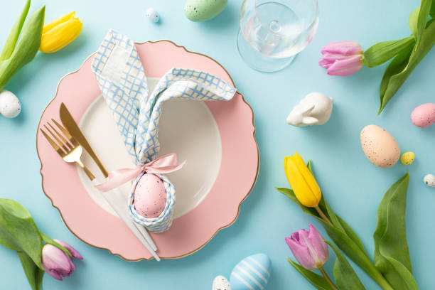 istock Celebrate Easter in style: Top view of a table featuring bunny ears napkin, small rabbit, cutlery, wine glass, tulips, sprinkles, and eggs. Pastel blue background adds a festive touch 2005516151