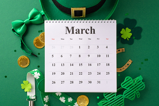 Celebrate St. Patrick's Day! Top view of a calendar, leprechaun hat, bow tie, suspenders, lucky horseshoe, party eyewear, and gold coins. Green background with trefoils, confetti, ready for notes
