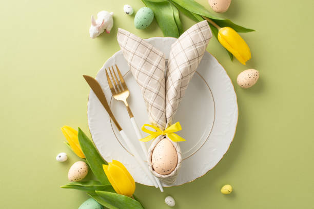 istock Easter-themed table setting: top view bunny-ear napkin on plate, polished cutlery, lively tulips, and colorful eggs on a pastel green surface. Ideal for a festive mood, text or promotions 2005516175