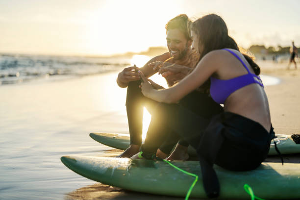 istock Couple sitting on surfboards and using phone while smiling at it. 2005529941