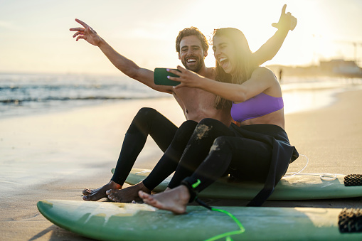 Cheerful couple is sitting on surfboards on a shore and taking selfies.